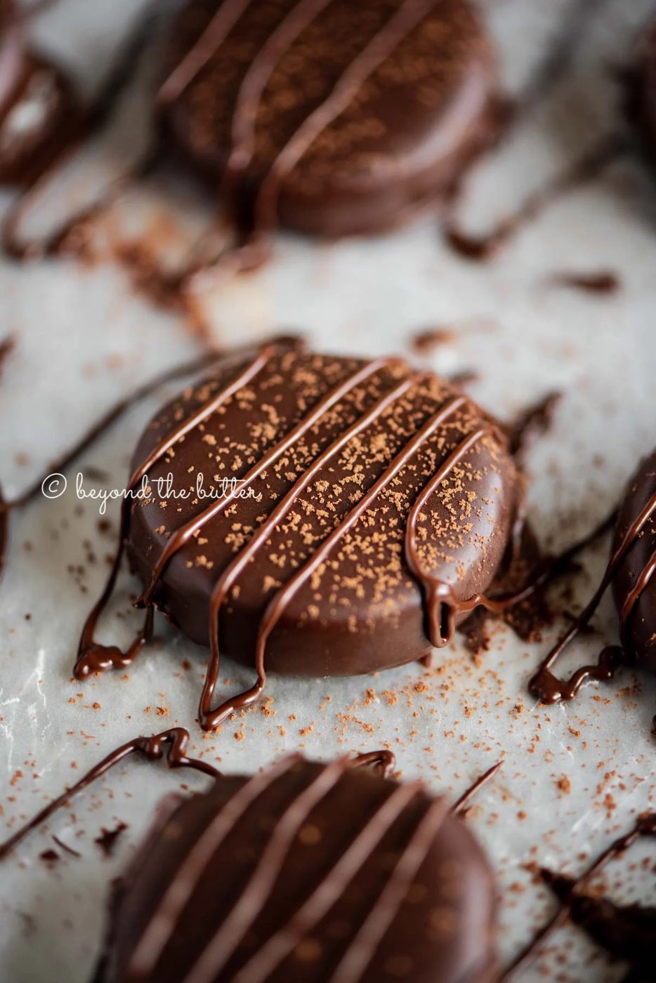 Round peppermint patties on a wax paper lined baking sheet sprinkled with cocoa powder and drizzled with melted chocolate.