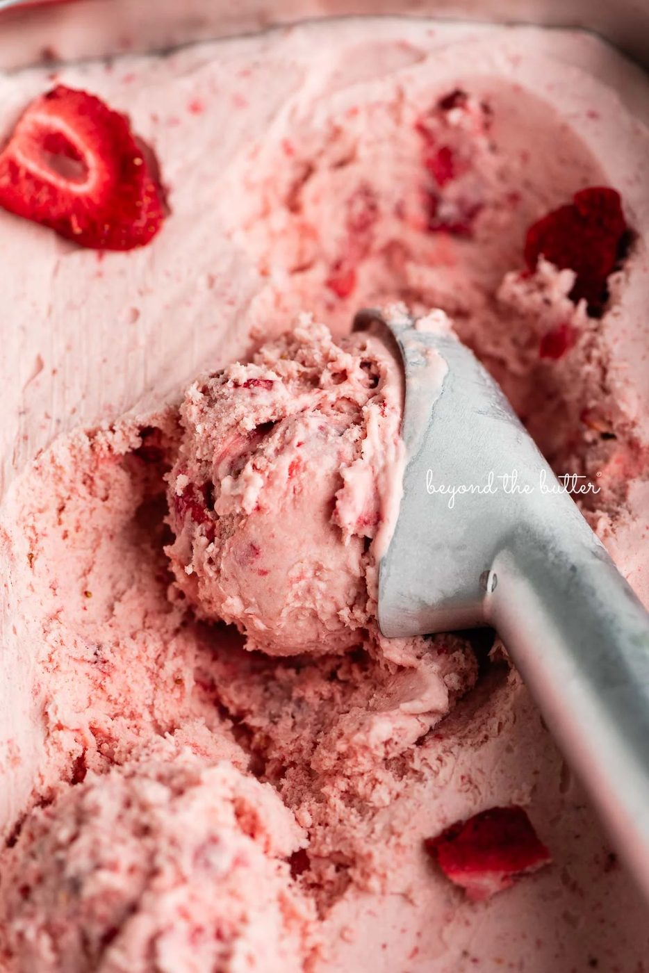 Container of strawberry ice cream with a scoop taken out using a stainless steel ice cream scoop.