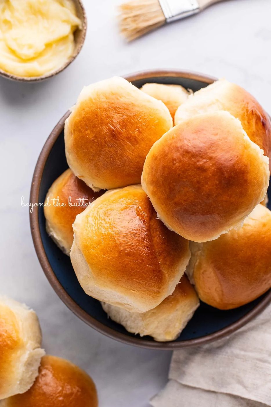 Blue ceramic bowl filled with house-made honey butter dinner rolls with a small bowl of honey butter and brush next to it on a white marbled background | © Sovuh