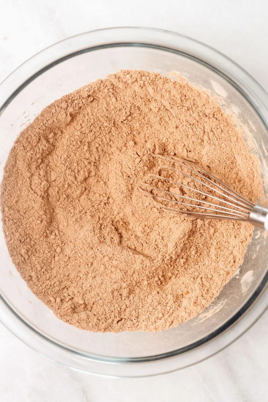 Sugar, flour, cocoa, baking powder, and salt whisked together in a large glass mixing bowl on a white marbled background.