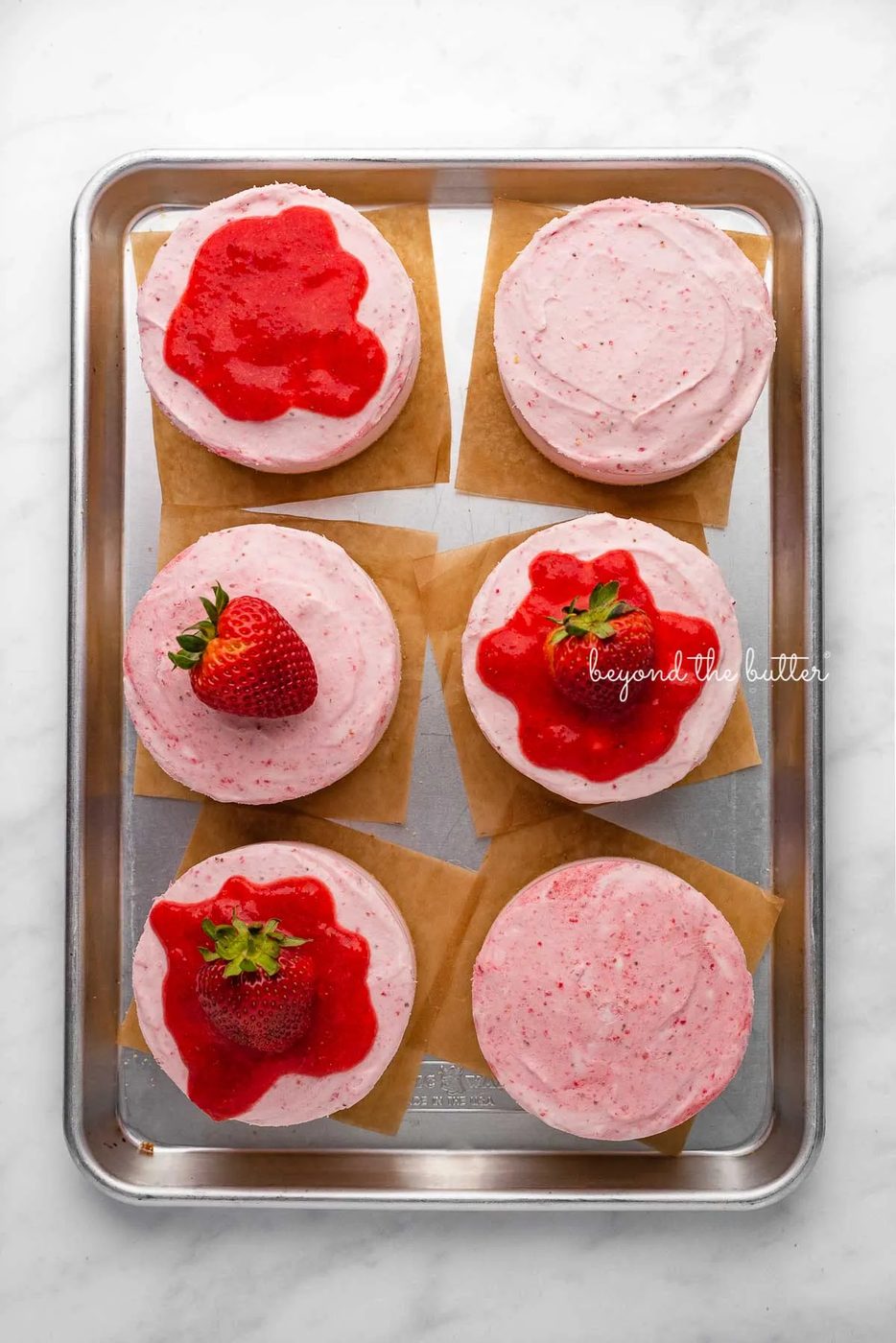 Individual no-bake strawberry cheesecakes topped with strawberry glaze and a fresh strawberry on a parchment paper-lined baking sheet | © Sovuh
