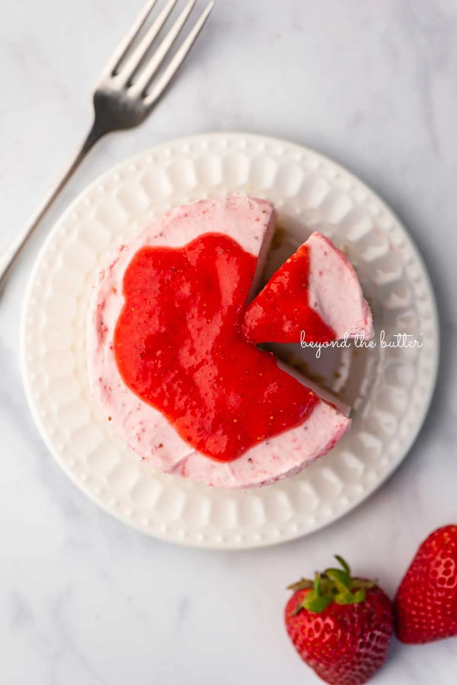 Small 4-inch no-bake strawberry cheesecake on a white dessert plate with a fork and fresh strawberries nearby on a marbled background | © Sovuh