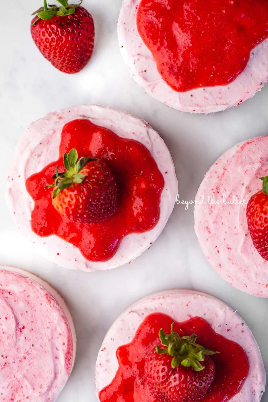 Small 4-inch no-bake strawberry cheesecakes topped with strawberry glaze and a fresh strawberry on a parchment paper-lined baking sheet | © Sovuh