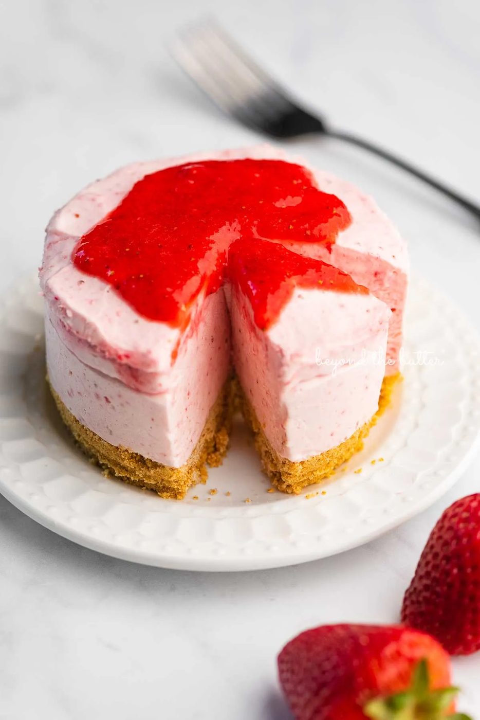 Small 4-inch no-bake strawberry cheesecake on a white dessert plate with a fork and fresh strawberries nearby on a marbled background | © Sovuh
