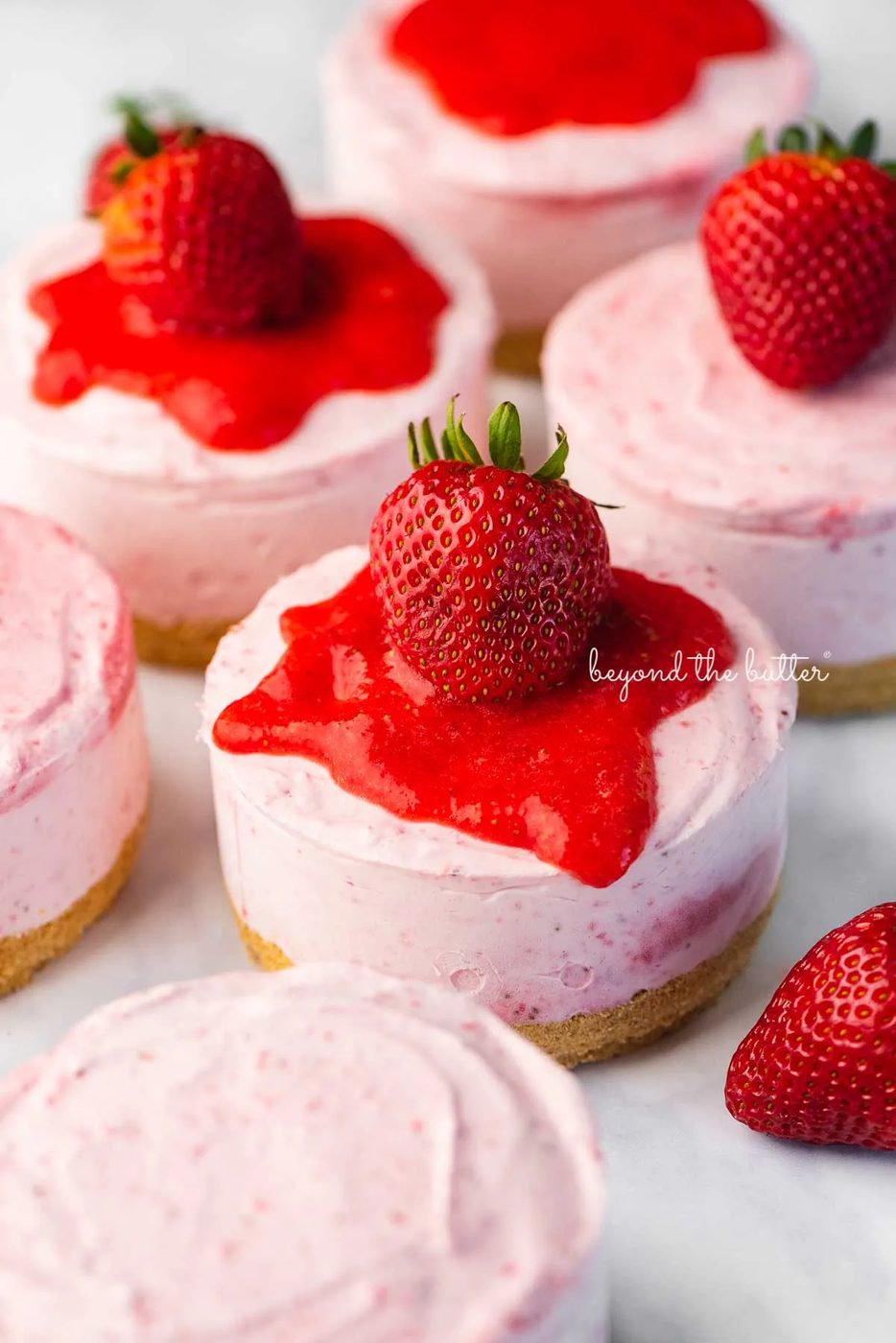 Individual no-bake strawberry cheesecakes topped with strawberry glaze and a fresh strawberry on a parchment paper-lined baking sheet | © Sovuh