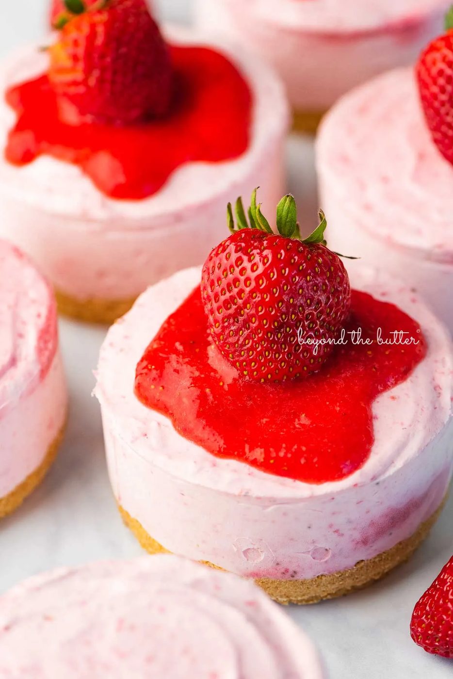 Individual no-bake strawberry cheesecakes topped with strawberry glaze and a fresh strawberry on a parchment paper-lined baking sheet | © Sovuh