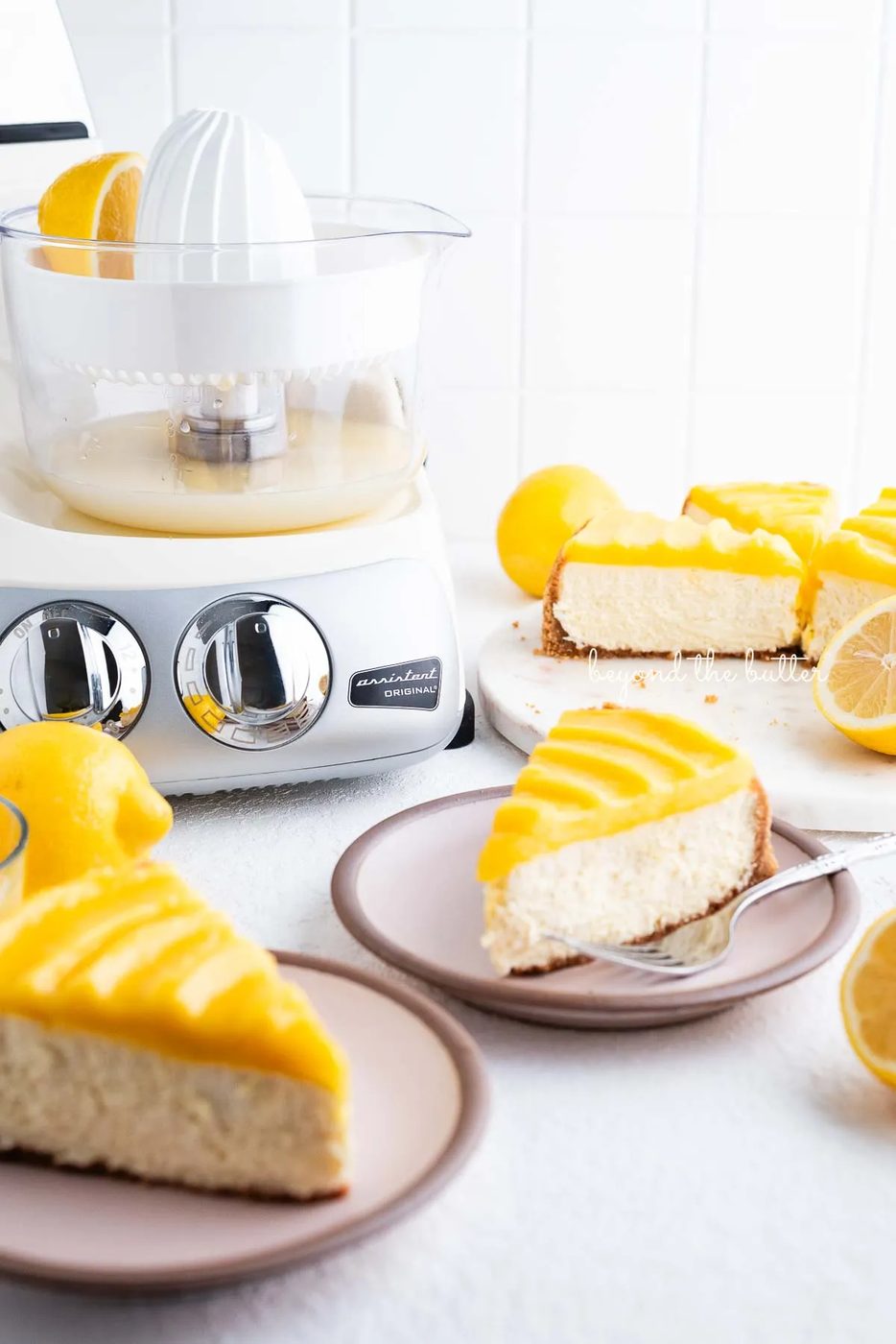 Slices of lemon cheesecake topped with lemon curd on pink dessert plates with cream colored Ankarsrum mixer on white background.