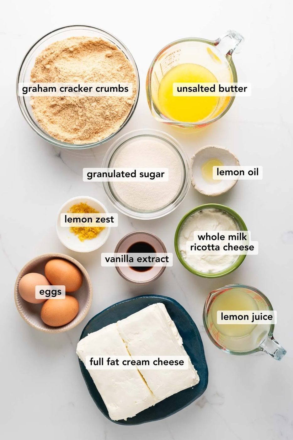 Labeled ingredients in bowls used to make lemon cheesecake on a white marbled background.