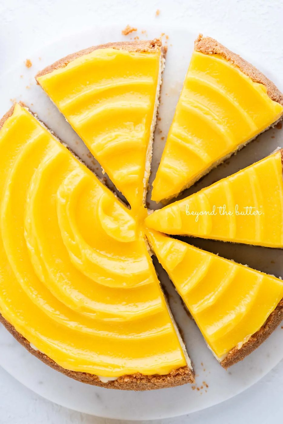 Lemon cheesecake topped with lemon curd cut into slices on a marble cake plate with white background.