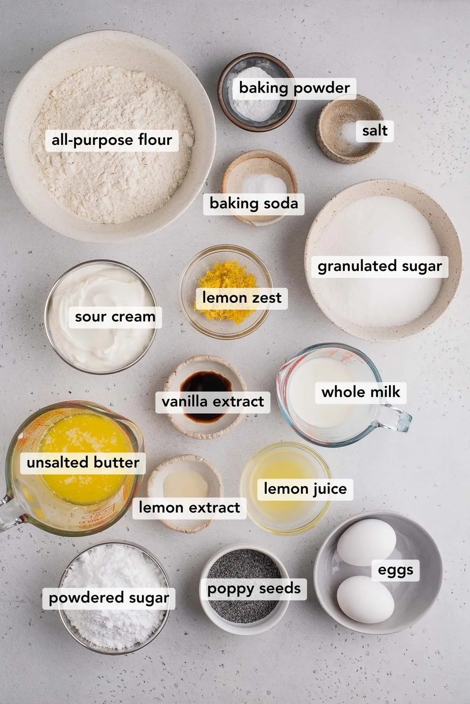 Bowls of different ingredients to make lemon poppy seed muffins on light gray background.