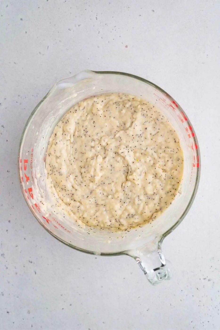 Melted butter and poppy seeds stirred into the lemon muffin batter in a large glass measuring bowl on a light grey background.