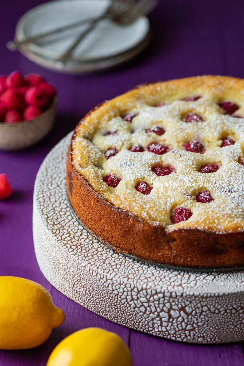 Lemon raspberry ricotta cake dusted with powdered sugar on crackled ceramic cake stand and purple wood background.