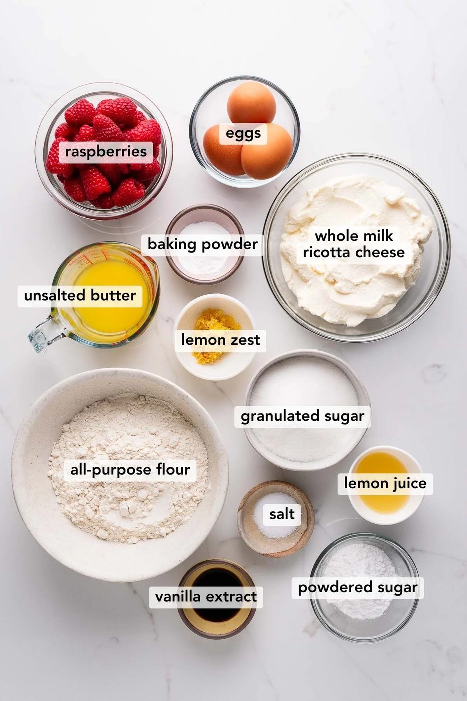 Labeled ingredients in bowls used to make lemon raspberry ricotta cake on a white marbled background.