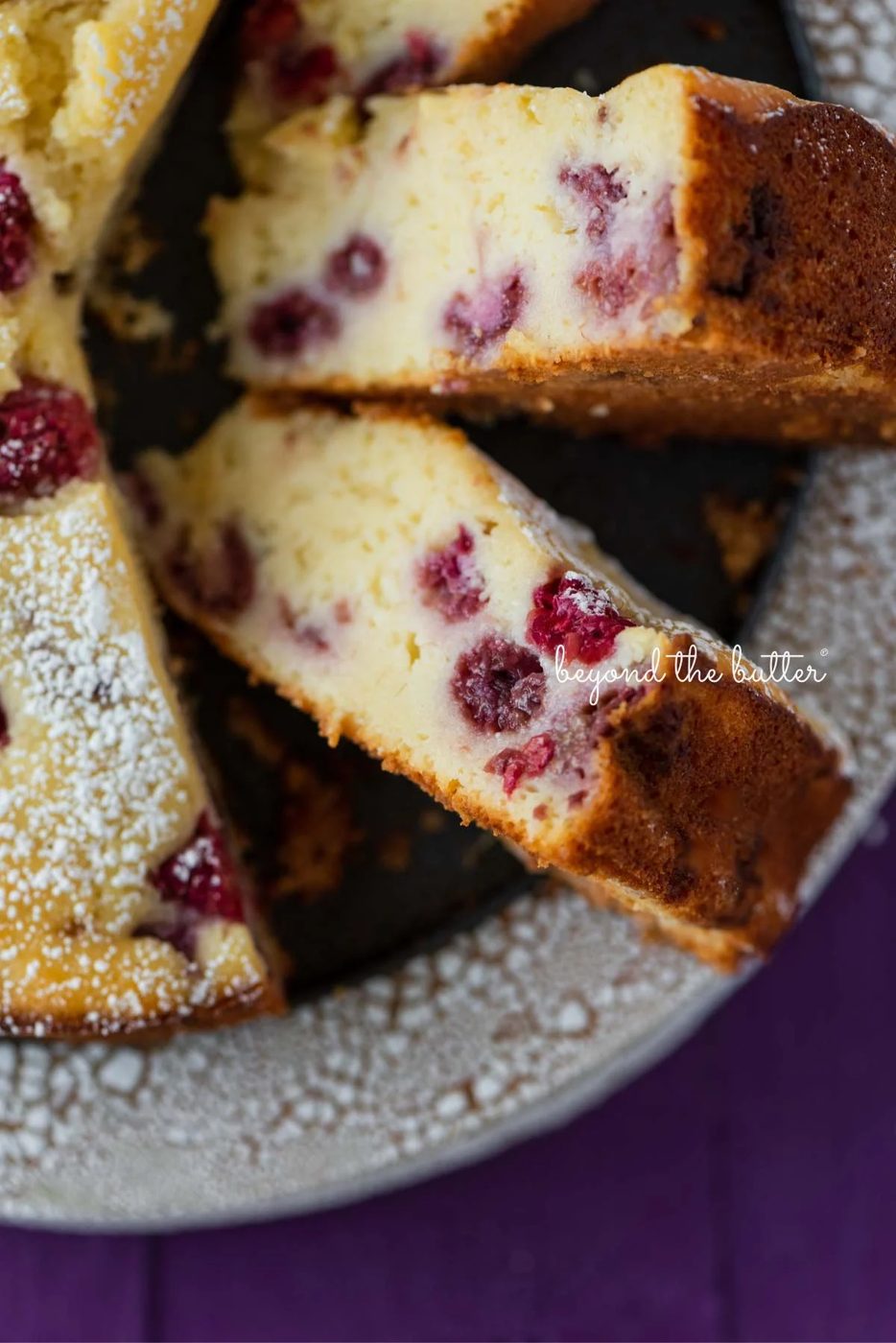 Lemon raspberry ricotta cake dusted with powdered sugar on crackled ceramic cake stand and purple wood background.