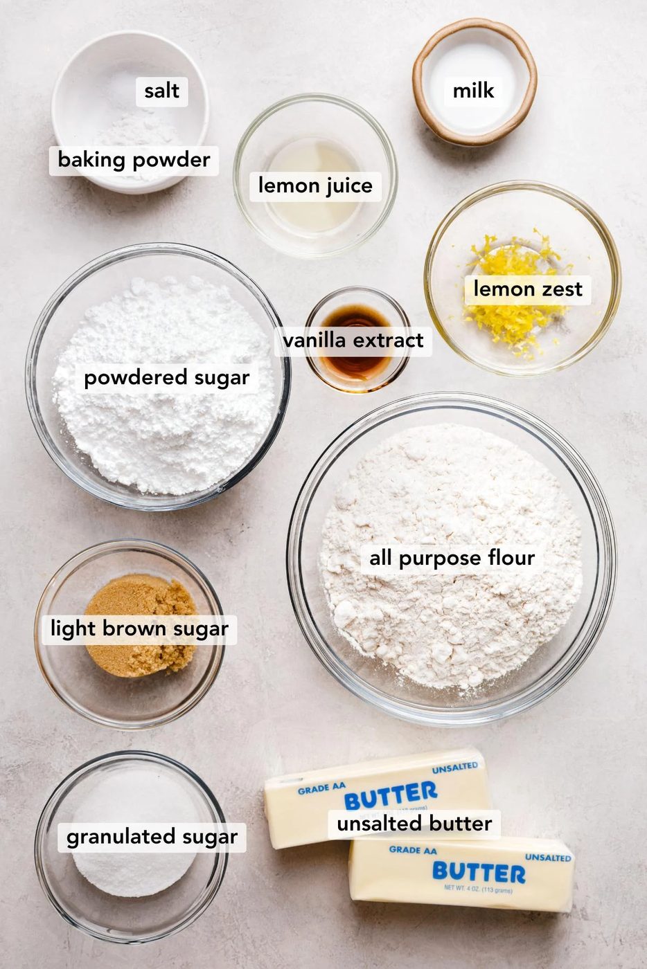 Ingredients in various size bowls labeled as salt, baking powder, milk, powdered sugar, vanilla extract, lemon juice, lemon zest, light brown sugar, all purpose flour, granulated sugar, and unsalted butter on a white marbled background.