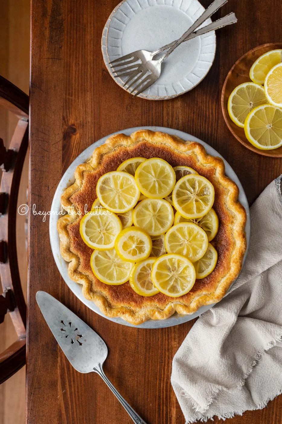 lemon sponge pie decorated with candied lemon sliced on a dark wood background with small dessert plates, napking, pie server, and small plate of sliced lemons | All images © Sovuh
