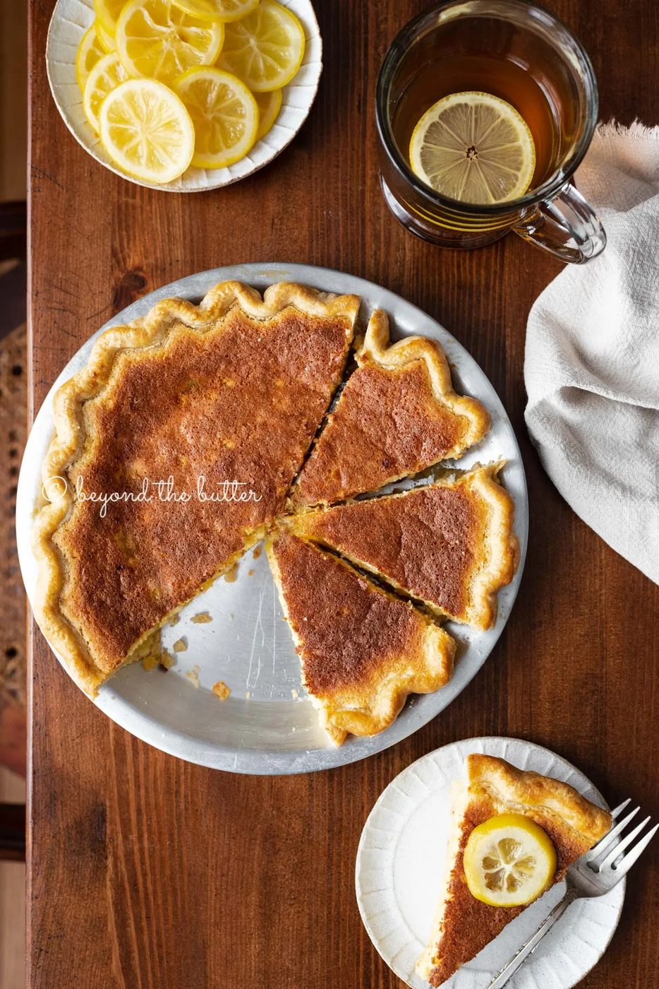 Lemon sponge pie with cut slices on dark wood background with hot lemon tea, small plate of sliced lemons, and slice to the side | All images © Sovuh
