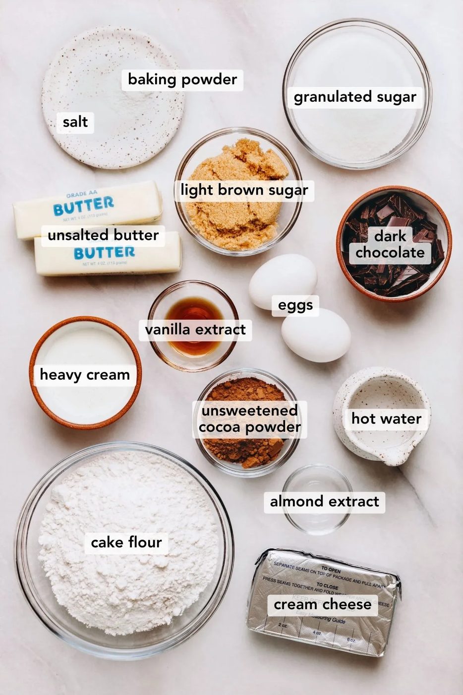 Ingredients in various size bowls and plates labeled as salt, baking powder, granulated sugar, light brown sugar, unsalted butter, heavy cream, vanilla extract, almond extract, cream cheese, hot water, unsweetened cocoa powder, cake flour, dark chocolate, and eggs on a white marbled background.