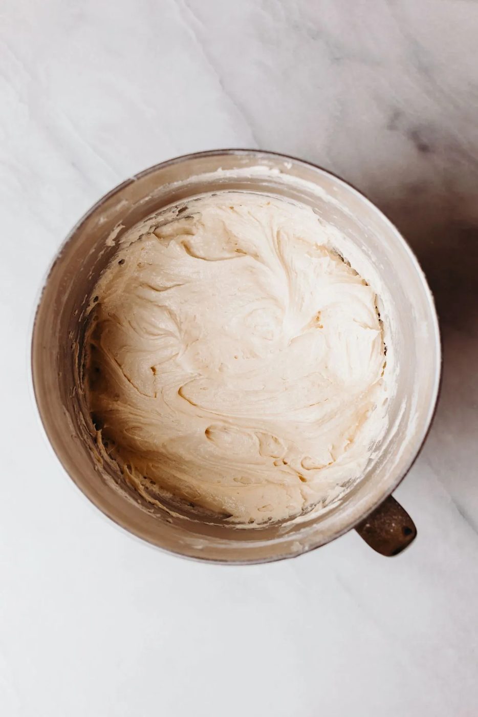 Bundt cake batter after adding the eggs and flour in a stainless steel mixing bowl on a white marbled background.