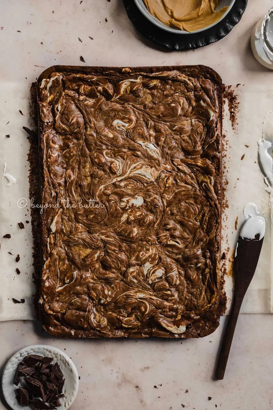 Uncut marshmallow peanut butter brownies on parchment paper and bowl of marshmallow fluff and spreader tool beside it | All Images © Sovuh