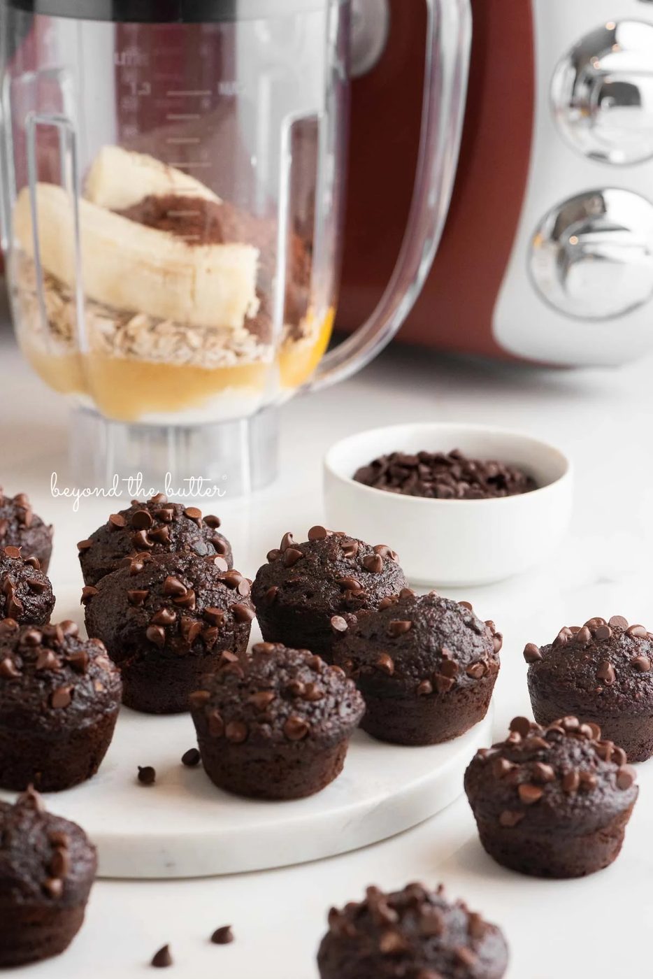 Mini chocolate banana oatmeal muffins placed in front of and around a rustic maroon colored Ankarsrum mixer and blender with small bowl of mini chocolate chips on a white background.