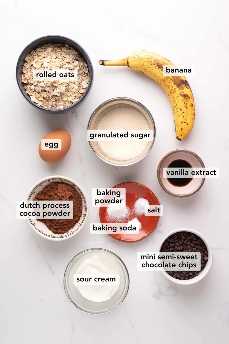 Ingredients in various size bowls labeled as rolled oats, banana, egg, granulated sugar, vanilla extract, dutch process cocoa powder, baking powder, salt, baking soda, mini semi-sweet chocolate chips, and sour cream on a white marbled background.