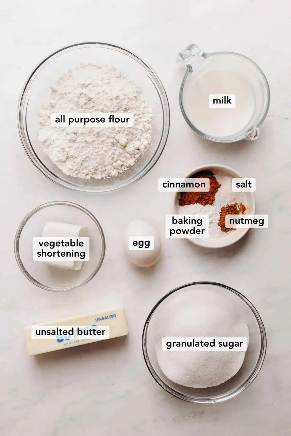 Ingredients in various size bowls labeled as all purpose flour, milk, vegetable shortening, cinnamon, salt, baking powder, nutmeg, egg, unsalted butter, and granulated sugar on a white marbled background.