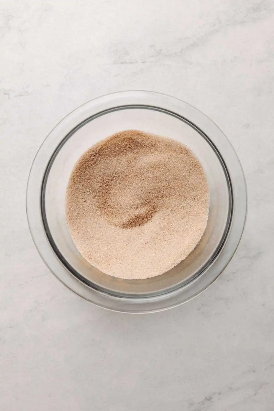 Cinnamon and sugar whisked together in small glass mixing bowl on a white marbled background.