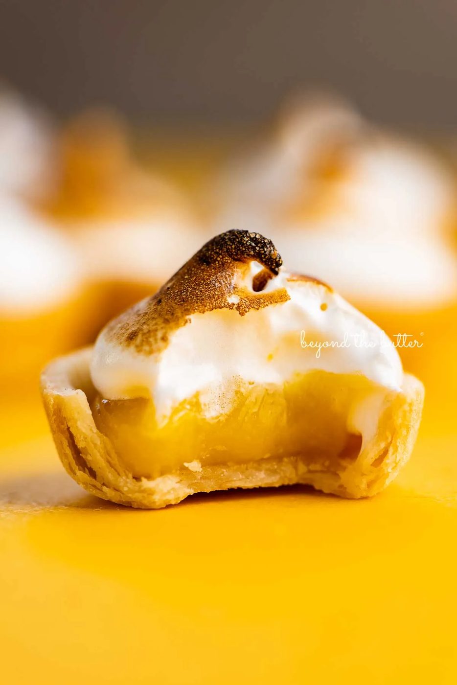 Mini lemon meringue pie with bite taken out surrounded by other mini pies on a yellow background | © Sovuh@