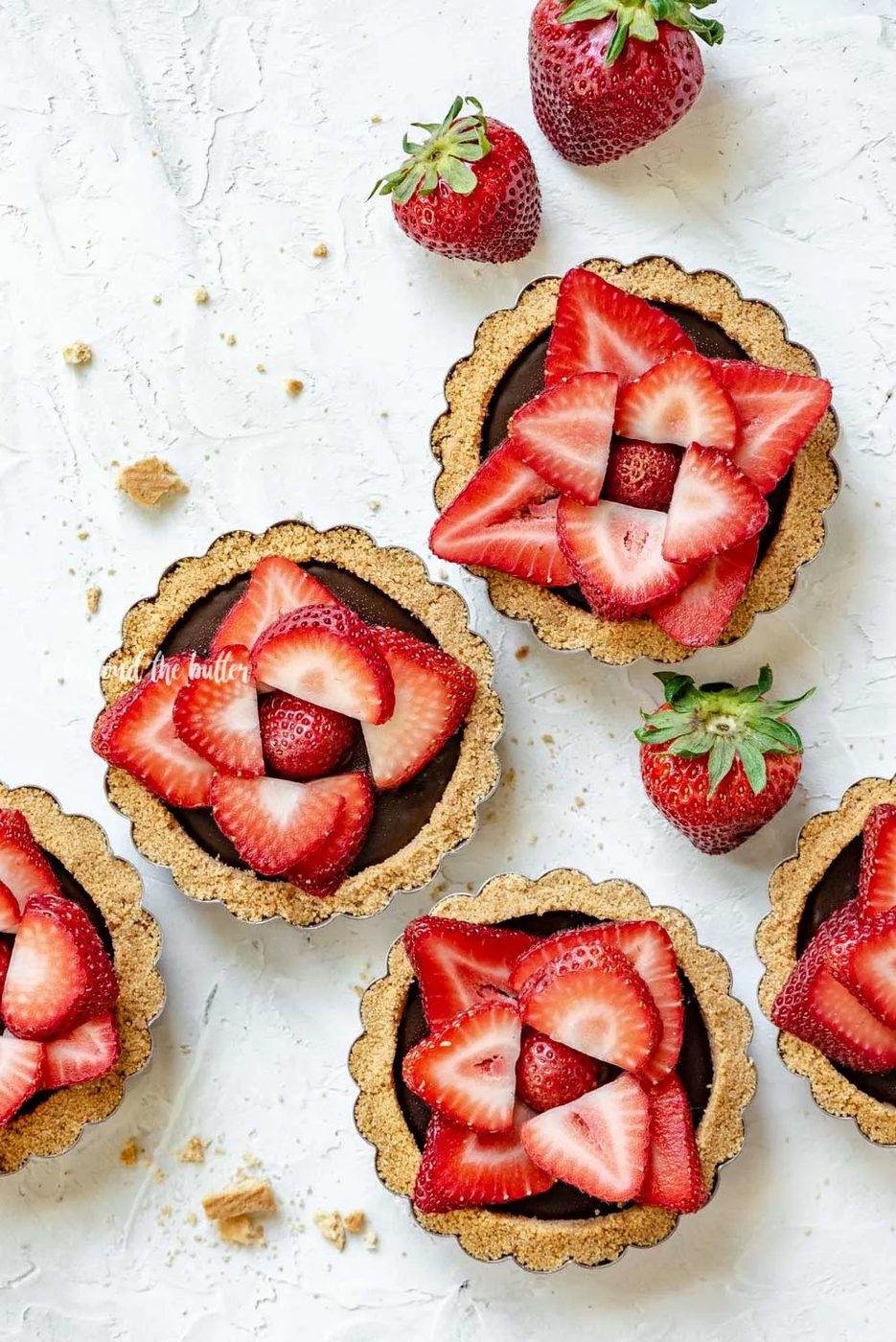 Overhead image of 6 mini strawberry nutella tarts on white stucco background with randomly placed strawberries around them | All Images © Sovuh