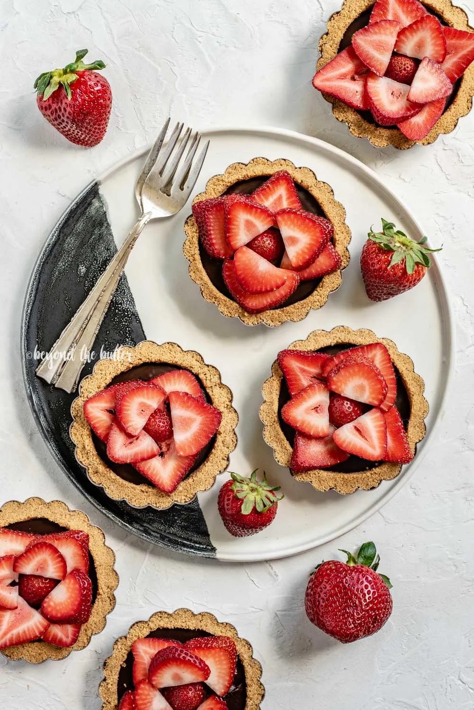 Overhead image of 6 mini strawberry nutella tarts on a black and white serving plate and randomly placed strawberries | All Images © Sovuh