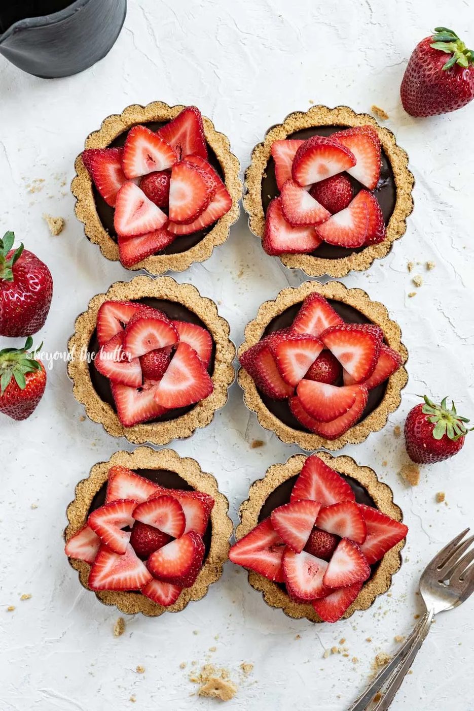 Overhead image of 6 mini strawberry nutella tarts on white stucco background with strawberries and forks around them | All Images © Sovuh