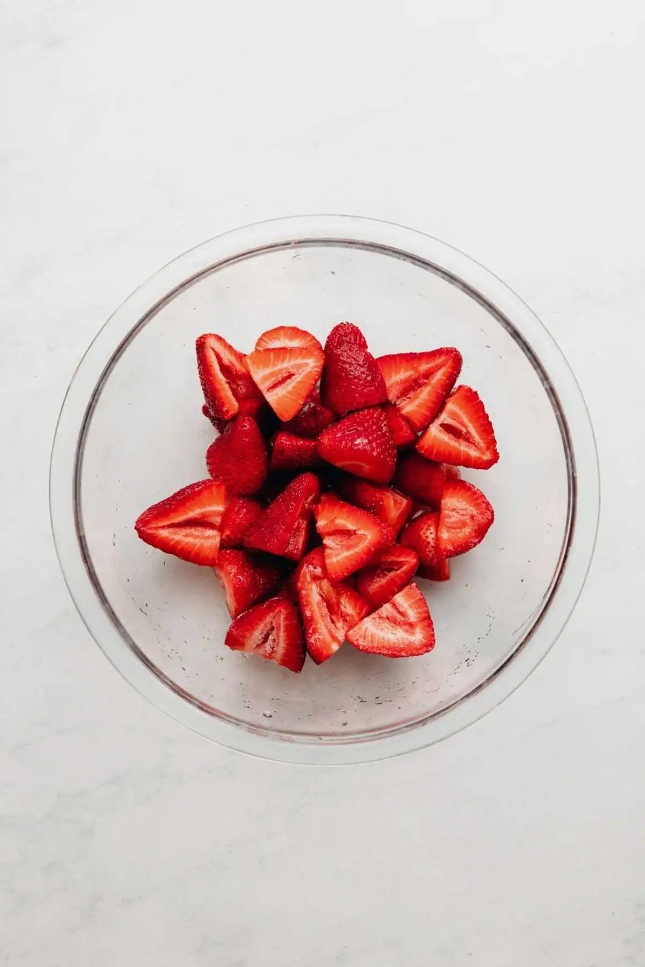 Sliced strawberries, lemon juice, and granulated sugar tossed together in a small glass bowl on a white and grey textured background.