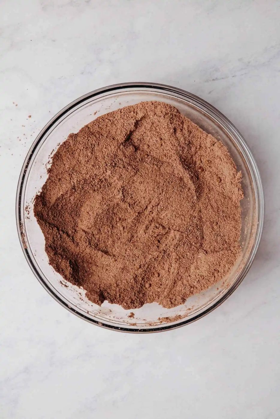 Whisked all purpose flour, granulated sugar, light brown sugar, dutch process cocoa powder, unsweetened cocoa powder, baking powder, baking soda, and salt in a medium size glass mixing bowl on a grey marbled background.