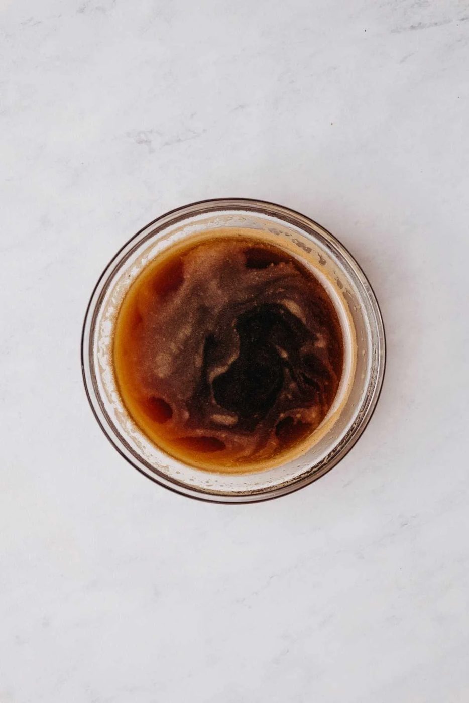 Espresso powder and hot water whisked together in a small glass mixing bowl on a grey marbled background.