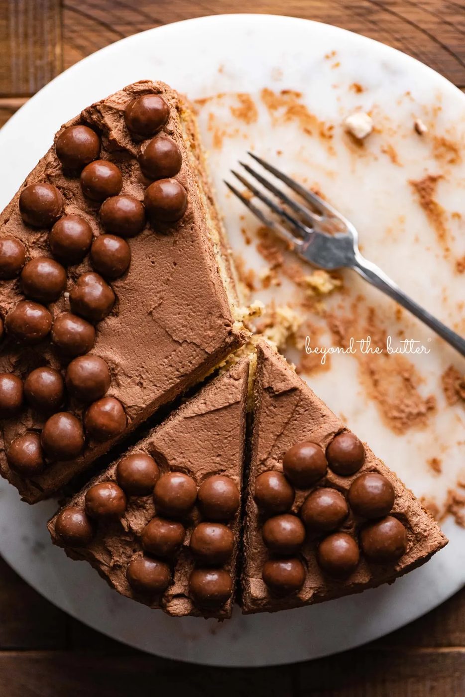 Top down view of banana cake frosted with malted chocolate mascarpone whipped cream and garnished with malted chocolate balls on marble cake board | © Sovuh