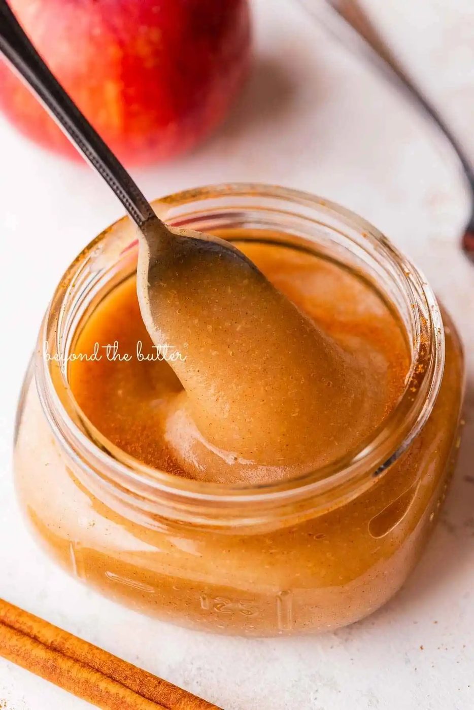 Filled glass jar of house-made applesauce with a spoon on white background with apples.