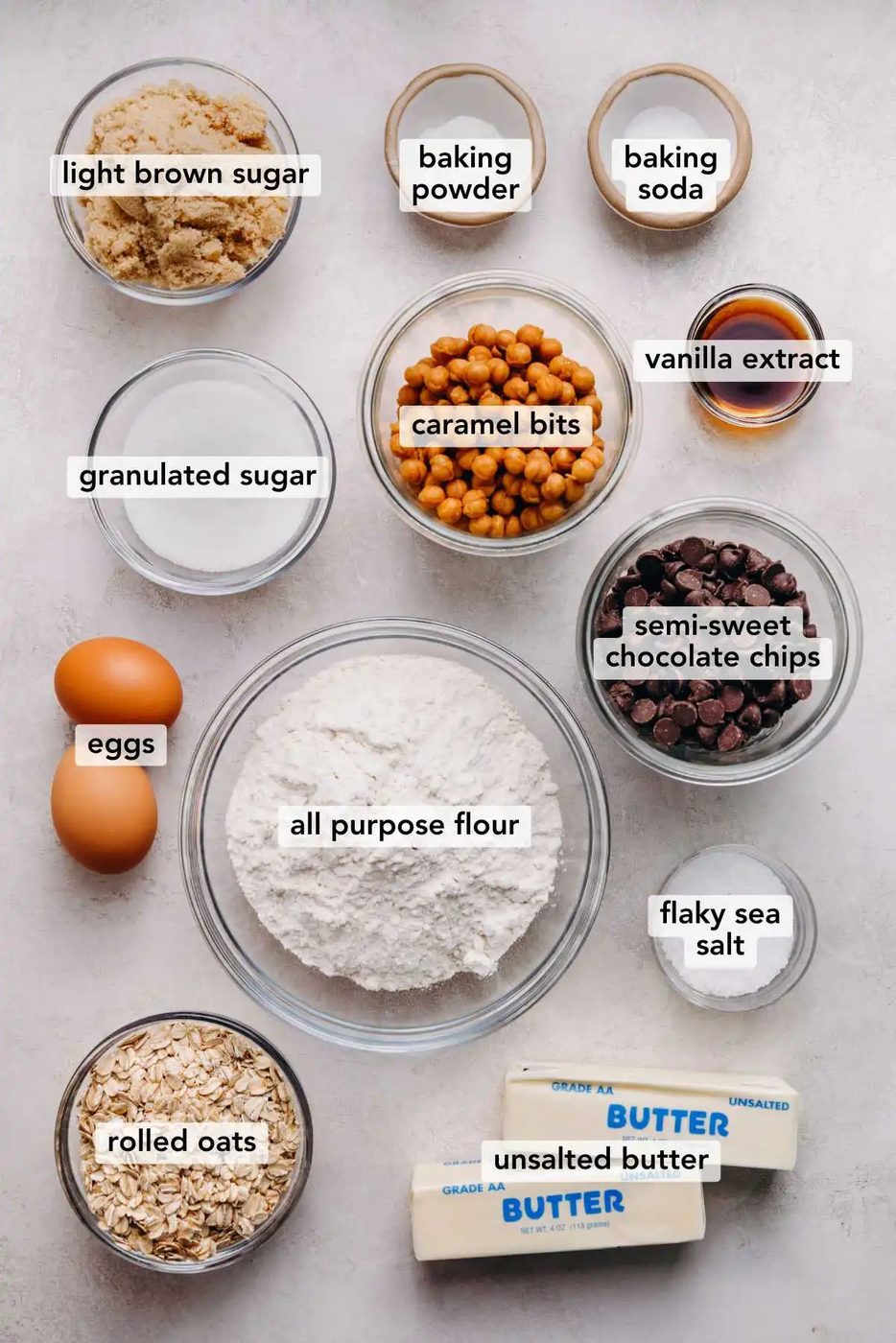 Ingredients in various size bowls labeled as light brown sugar, baking powder, baking soda, granulated sugar, caramel bits, vanilla extract, eggs, all purpose flour, semi sweet chocolate chips, rolled oats, flaky sea salt, and unsalted butter on a white marbled background.
