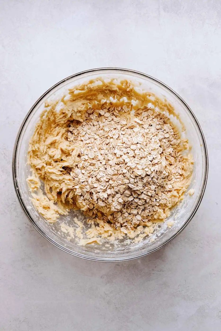 Rolled oats added to the top of oatmeal cookie dough mixture in a glass mixing bowl on a white grey marbled background.