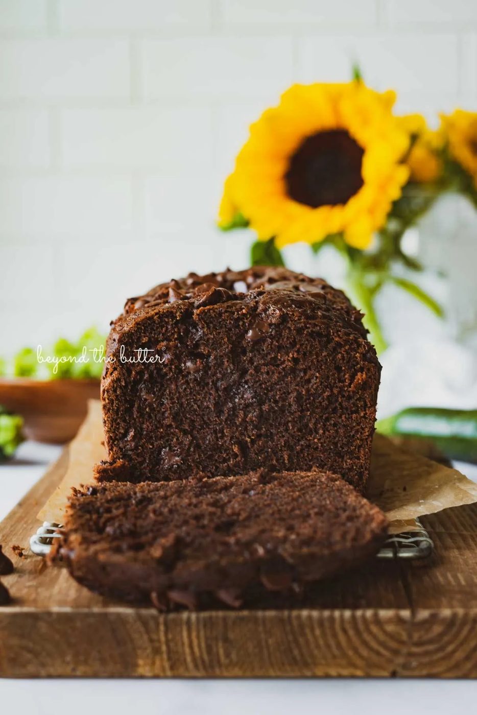 Sliced chocolate zucchini bread on a cutting board with white tile background and sunflowers | © Sovuh