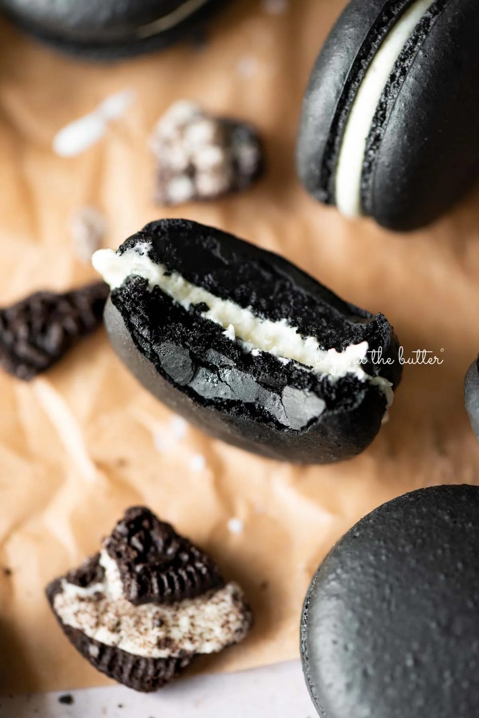 Half eaten oreo macaron on parchment paper surrounded by oreo macarons and broken pieces of oreo cookies.