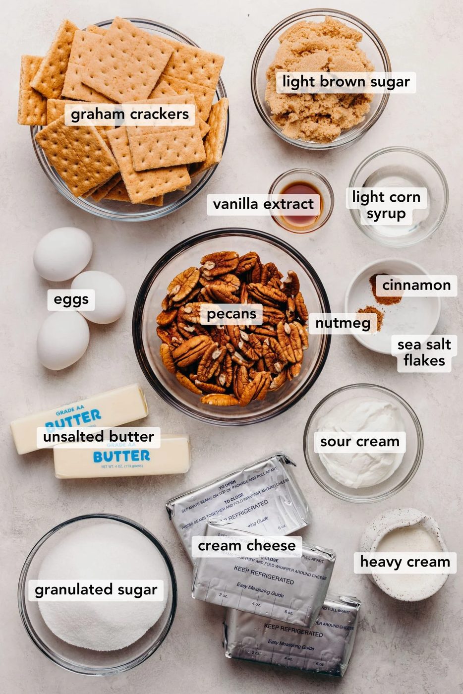 Ingredients in various size bowls and plates labeled as graham crackers, light brown sugar, vanilla extract, light corn syrup, eggs, pecans, nutmeg, cinnamon, sea salt flakes, unsalted butter, sour cream, heavy cream, granulated sugar, and cream cheese on a white marbled background.