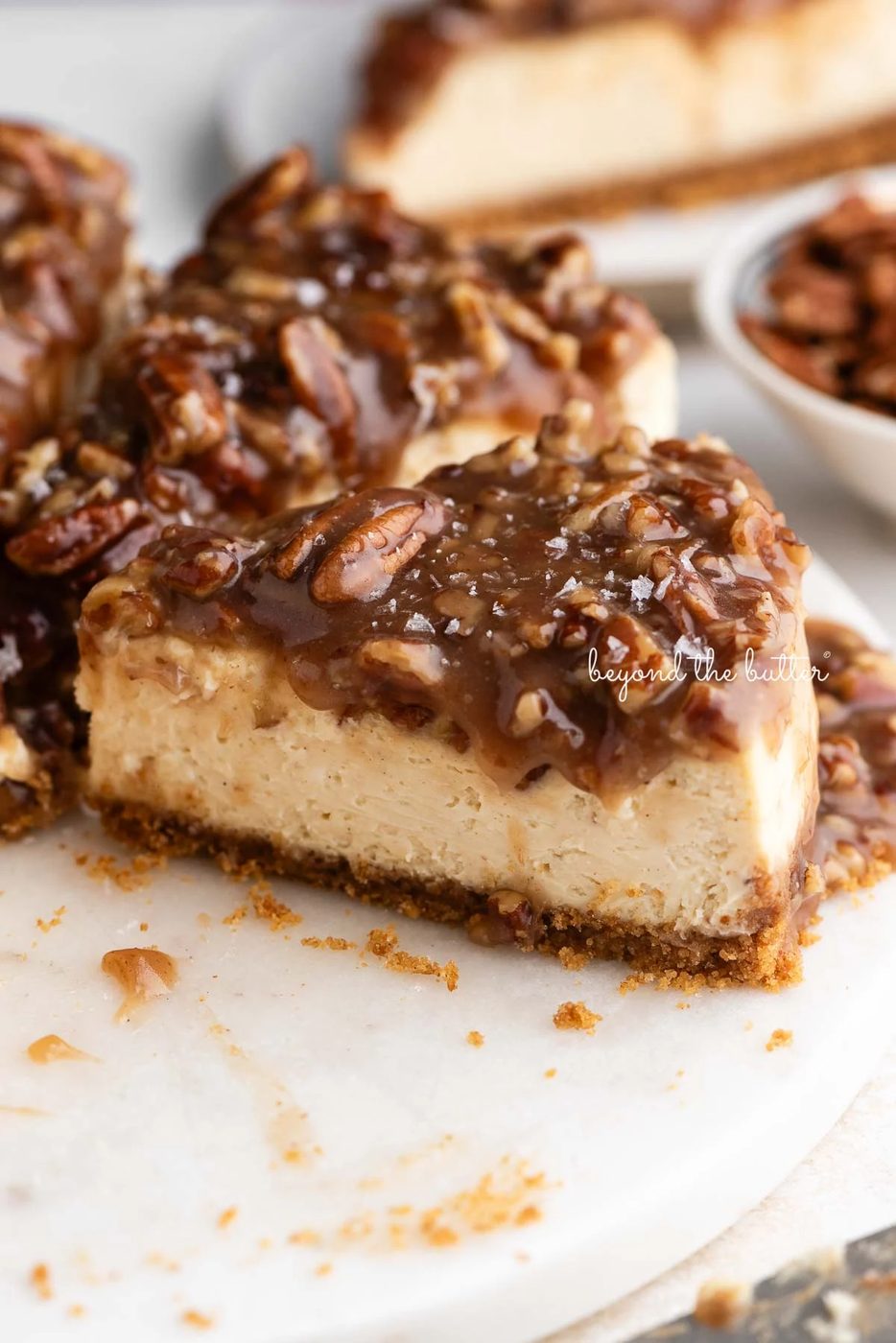 Sliced pecan pie cheesecake sprinkled with sea salt flakes on a marble cake stand next to a small bowl of pecans on white background.