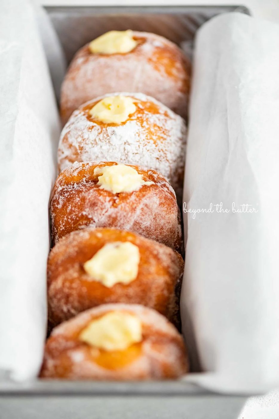 Single row of pennsylvania dutch fasnacht doughnuts filled with vanilla pastry cream wrapped in parchment paper in a loaf pan | © Sovuh
