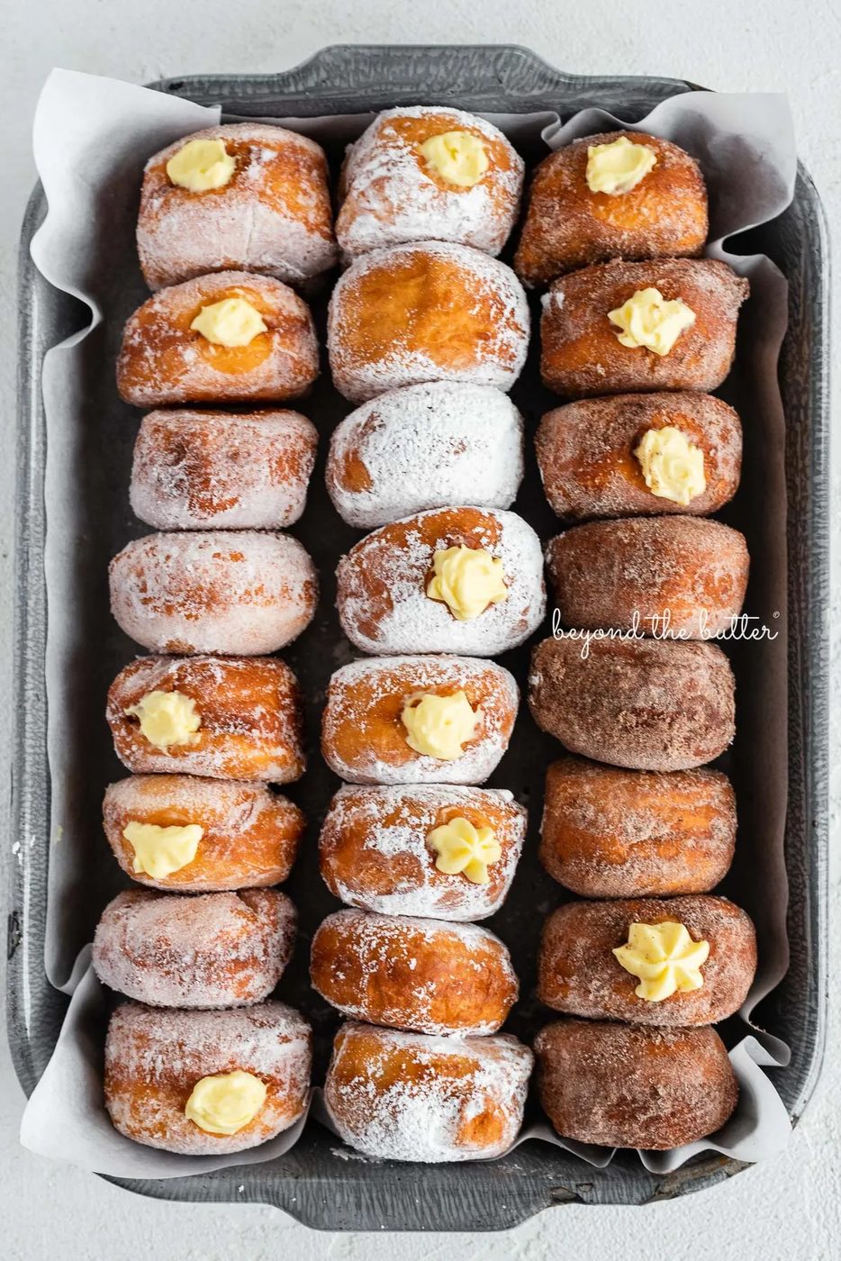 Rows of pennsylvania dutch fasnacht doughnuts in a parchment paper lined baking tray filled with pastry cream | © Sovuh