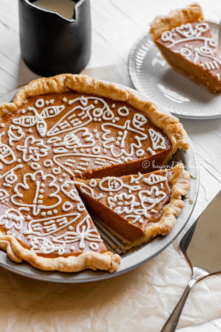 Angled image of cut pumpkin pie with a piped cream cheese frosting decoration surrounded by pie server, cinnamon sticks, plates, and small pitcher of milk | All images © Sovuh