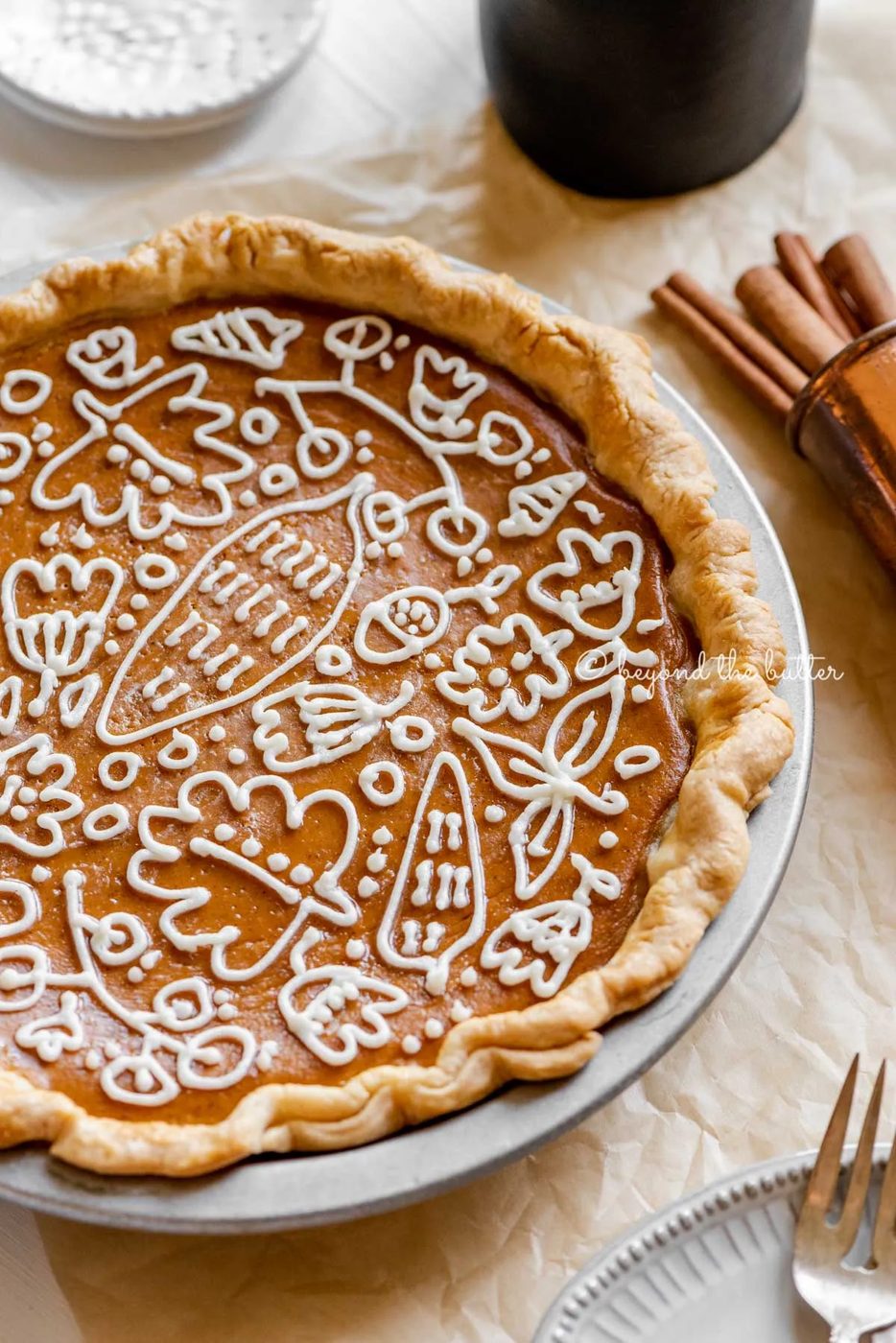 Angled image of pumpkin pie with a piped cream cheese frosting decoration surrounded by forks, cinnamon sticks, plates, and small pitcher of milk | All images © Sovuh