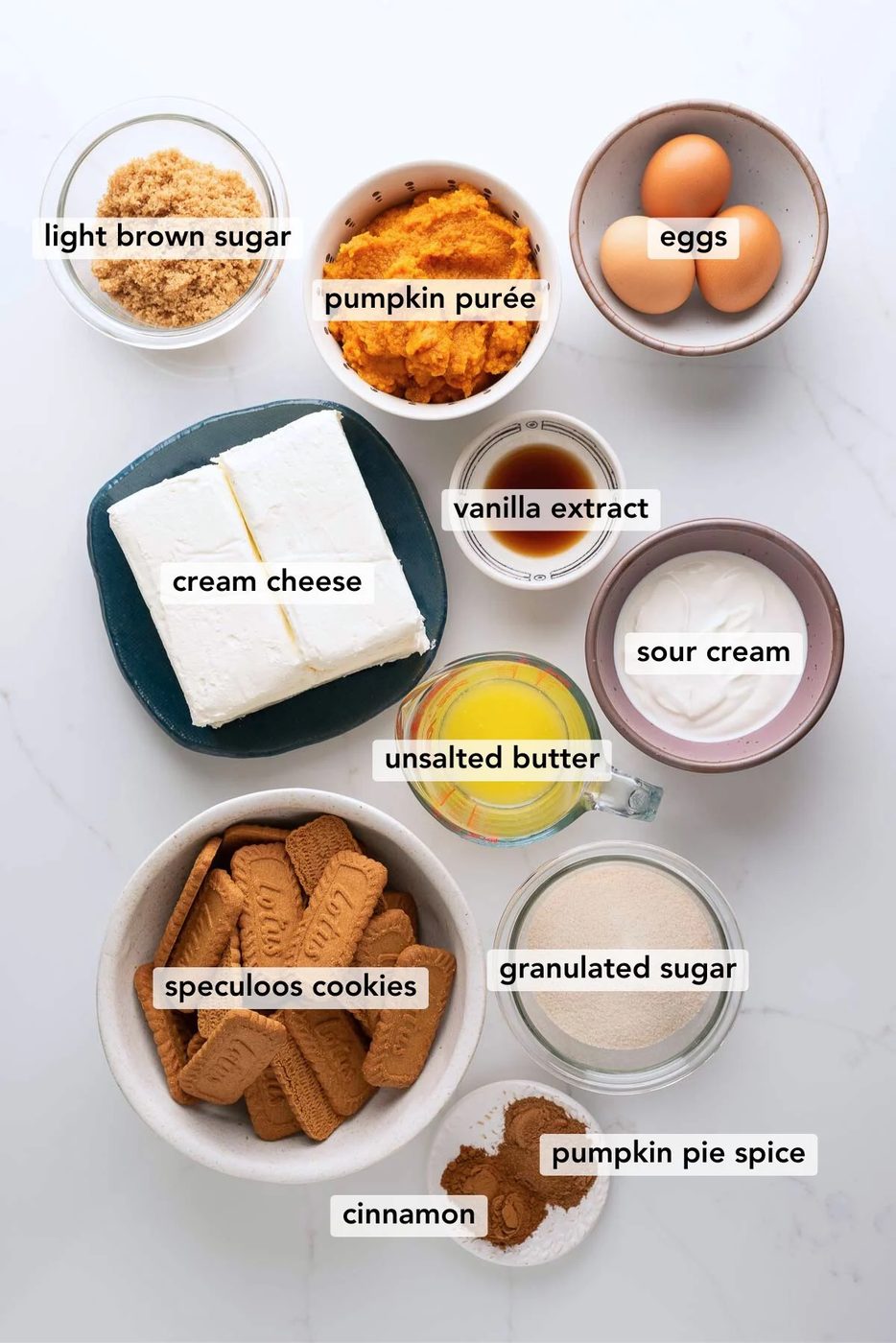 Ingredients in various size bowls labeled as light brown sugar, pumpkin puree, eggs, cream cheese, vanilla extract, unsalted butter, sour cream, speculoos cookies,granulated sugar, cinnamon, and pumpkin pie spice on a white marbled background.