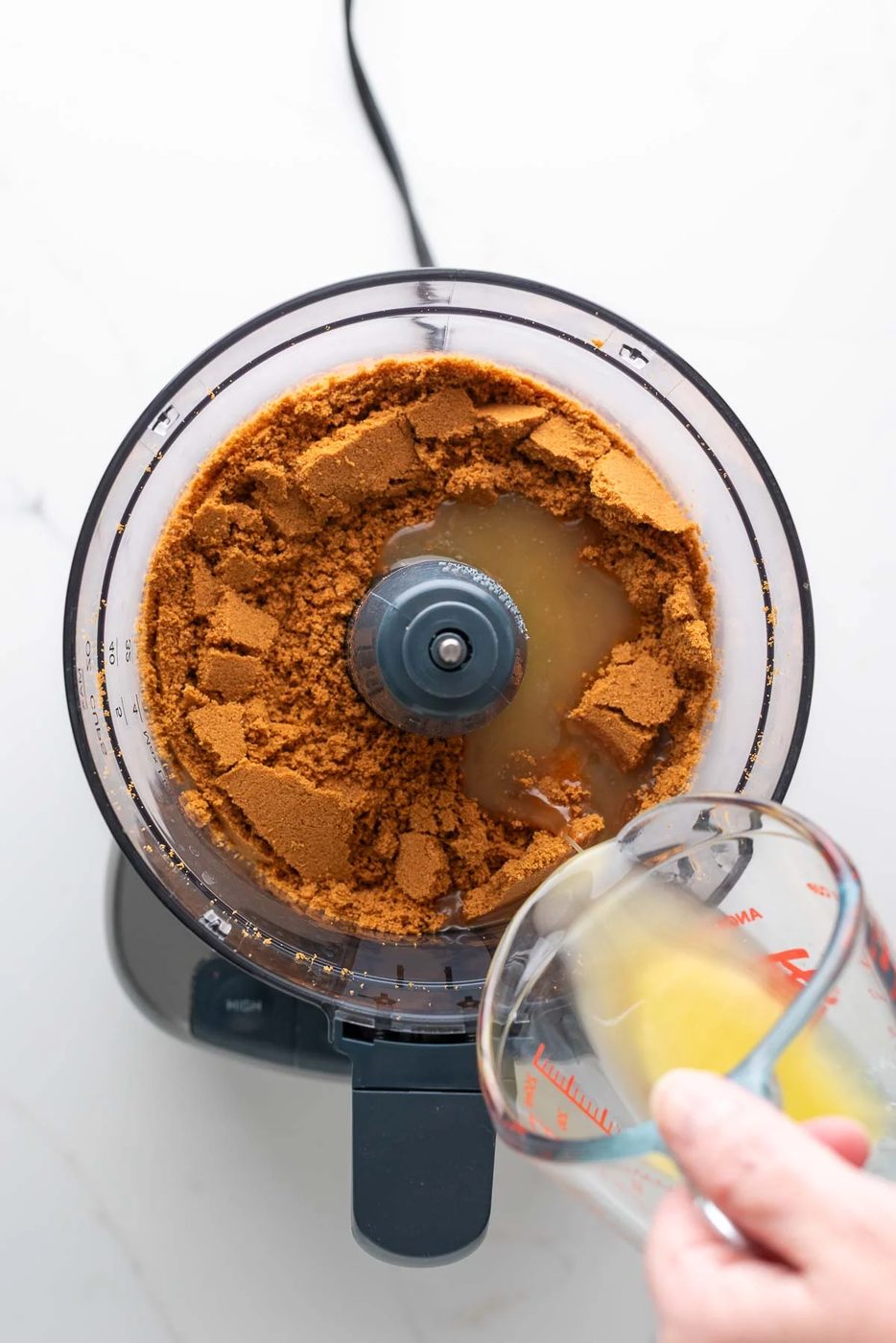 Adding melted unsalted butter in a glass measuring cup to a food processor with speculoos cookie crumbs.