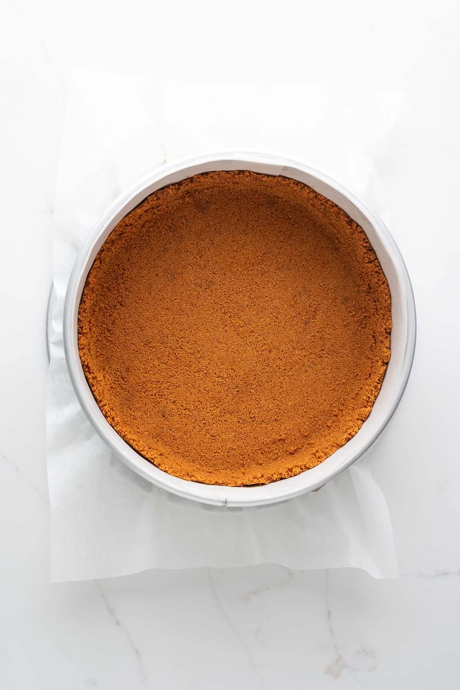 Baked speculoos cookie crust in a parchment paper lined springform pan on a white marbled background.